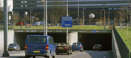 De Koningstunnel in Den Haag. Op de achtergrond zie je het centraal station. De bovenkant van de tunnel is bedekt met gras en bomen. De tunnel bestaat ui twee rijstroken in beide richtingen gescheiden door een verhoging waar lantaarnpalen staan.