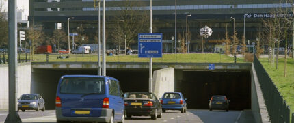 De Koningstunnel in Den Haag. Op de achtergrond zie je het centraal station. De bovenkant van de tunnel is bedekt met gras en bomen. De tunnel bestaat ui twee rijstroken in beide richtingen gescheiden door een verhoging waar lantaarnpalen staan.